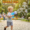 A young boy is joyfully firing a blue bubble gun, creating many bubbles in an outdoor park setting.