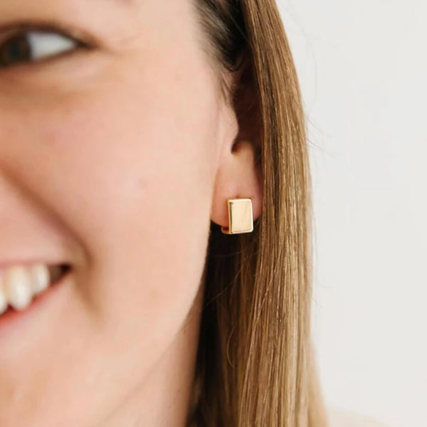 Primer plano de una mujer vistiendo aretes cuadrados geométricos dorados.