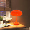 An orange mushroom-shaped LED table lamp with a slender stem sits on a wooden desk.
