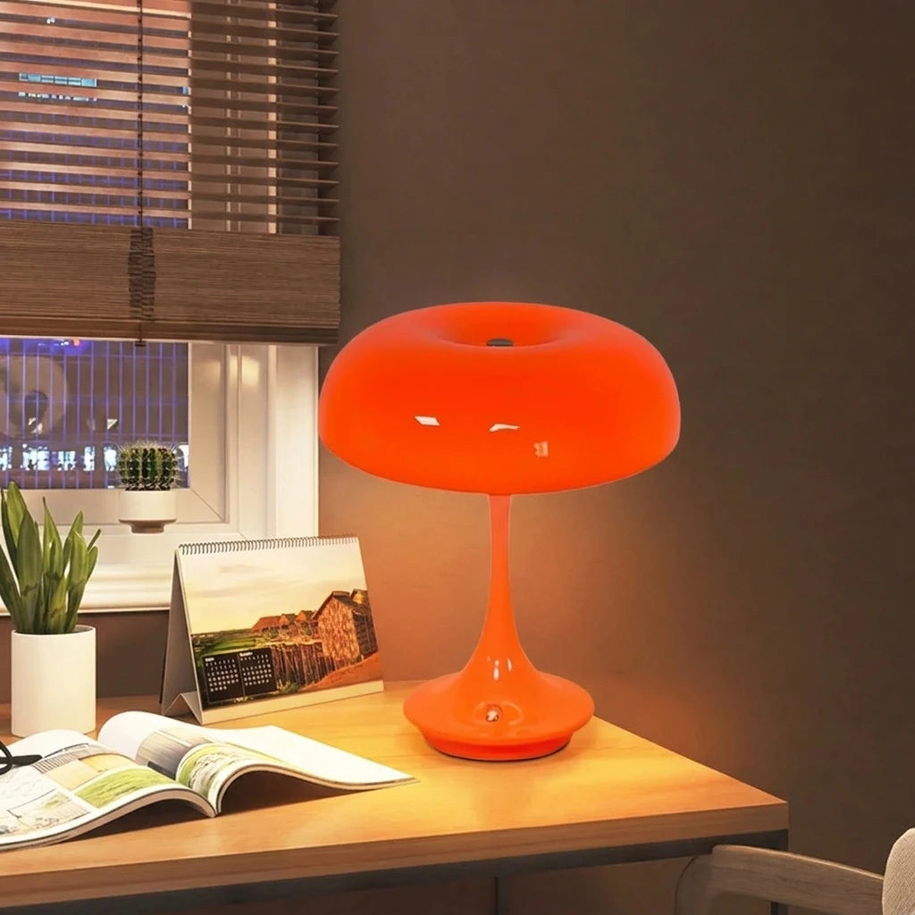 An orange mushroom-shaped LED table lamp with a slender stem sits on a wooden desk.