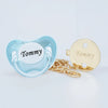 A light blue silicone pacifier and clip set with a white insert engraved with the name Tommy, accompanied by a gold chain.