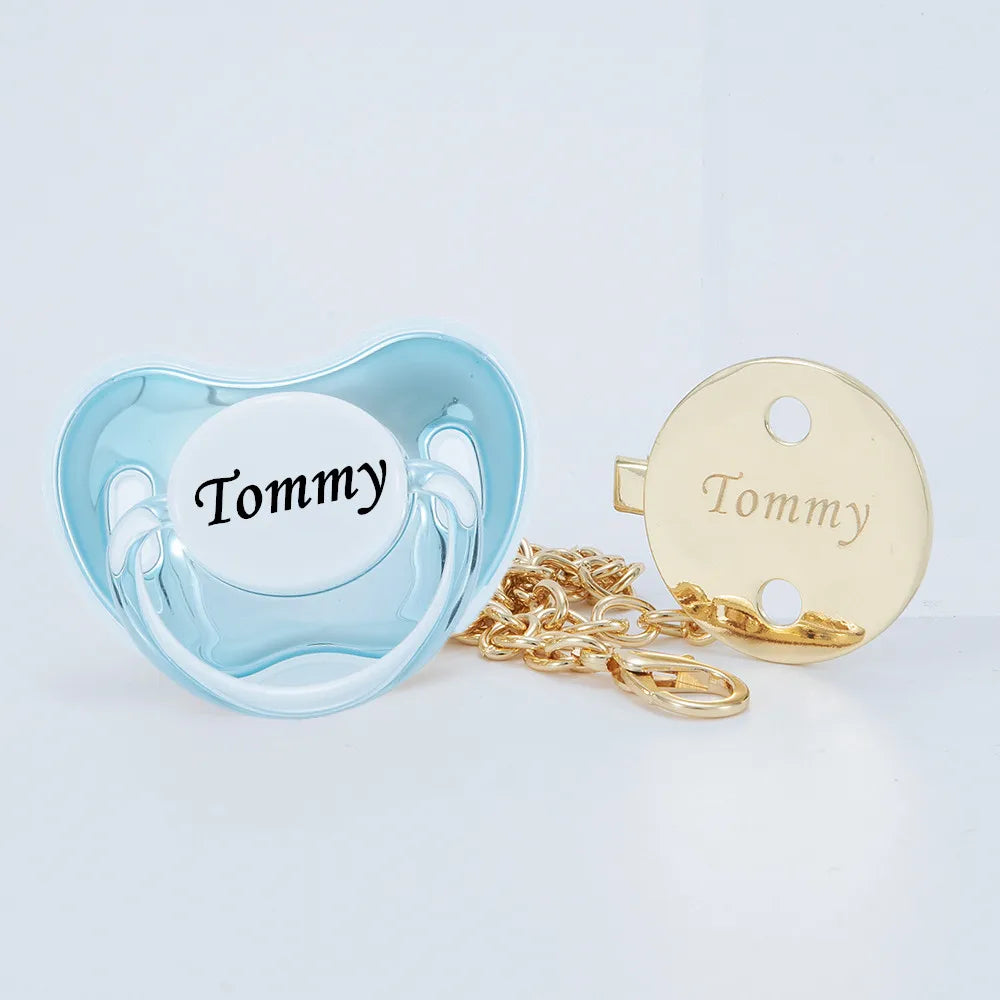 A light blue silicone pacifier and clip set with a white insert engraved with the name Tommy, accompanied by a gold chain.