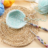 A crochet hook set with sunflower designs is shown next to a light blue crocheted bowl on a woven mat.