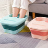 Two collapsible foot spa buckets in teal and pink resting on a gray rug next to a person's legs.