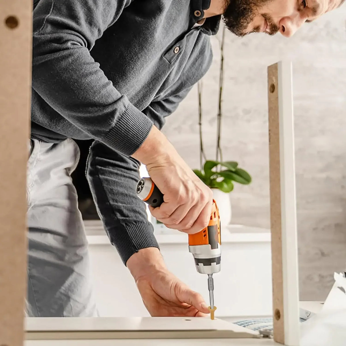 A person is using an orange power drill to drive a wood screw into a white wooden frame.