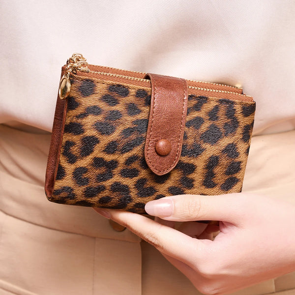 A brown leopard print tri-fold wallet with a leather flap and gold zipper is held in a hand.