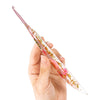 A hand holds a clear epoxy resin floral-patterned crochet hook set with pink and gold accents.