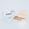 A clear silicone pacifier and a gold-toned clip with the name Tommy engraved on it.
