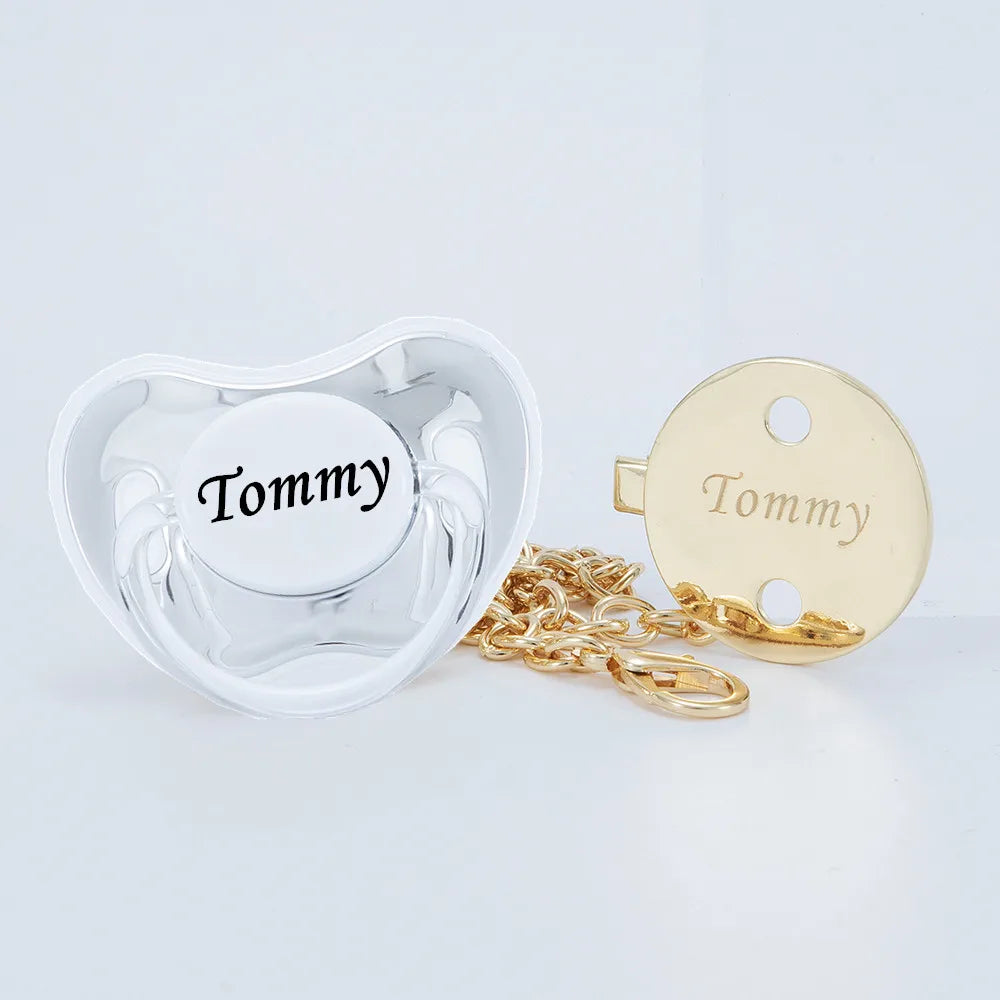 A clear silicone pacifier and a gold-toned clip with the name Tommy engraved on it.