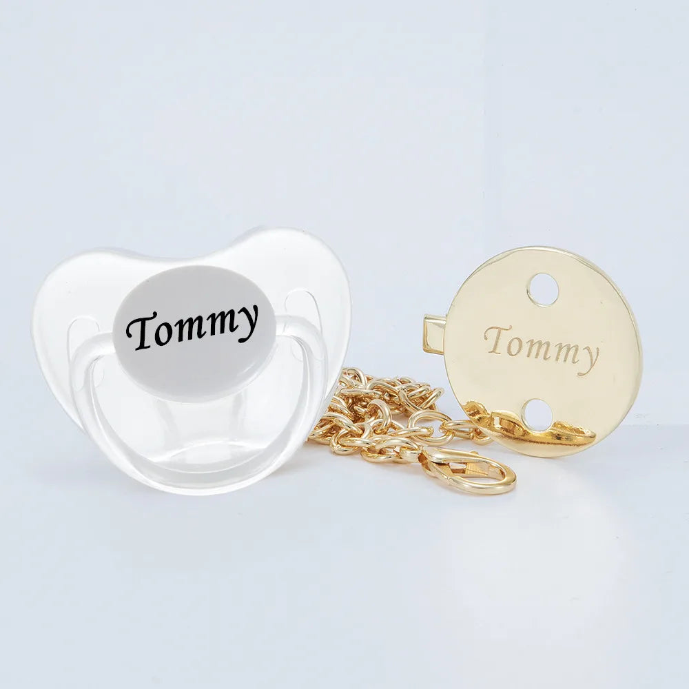 A clear silicone pacifier with the name Tommy and a gold-colored clip set.