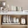 A white expandable pot and pan organizer rack with adjustable dividers is displayed on a wooden shelf.
