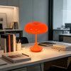 An orange mushroom-shaped LED table lamp with a slender stem sits on a white desk surface.