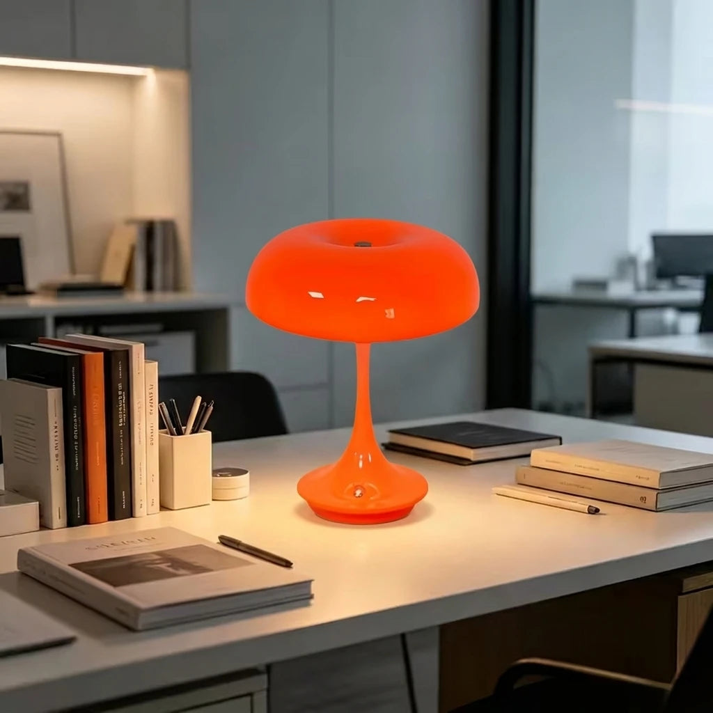 An orange mushroom-shaped LED table lamp with a slender stem sits on a white desk surface.