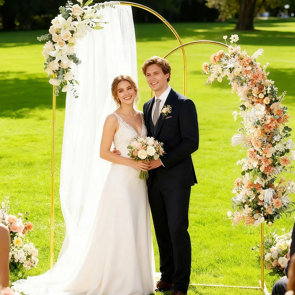 A bride and groom stand in front of a gold metal wedding arch backdrop with white draping and floral arrangements on a green 