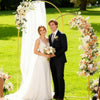 A bride and groom stand in front of a gold metal wedding arch backdrop with white draping and floral arrangements on a green 