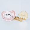 A pink silicone pacifier and a gold-tone clip with the name Tommy engraved on it.