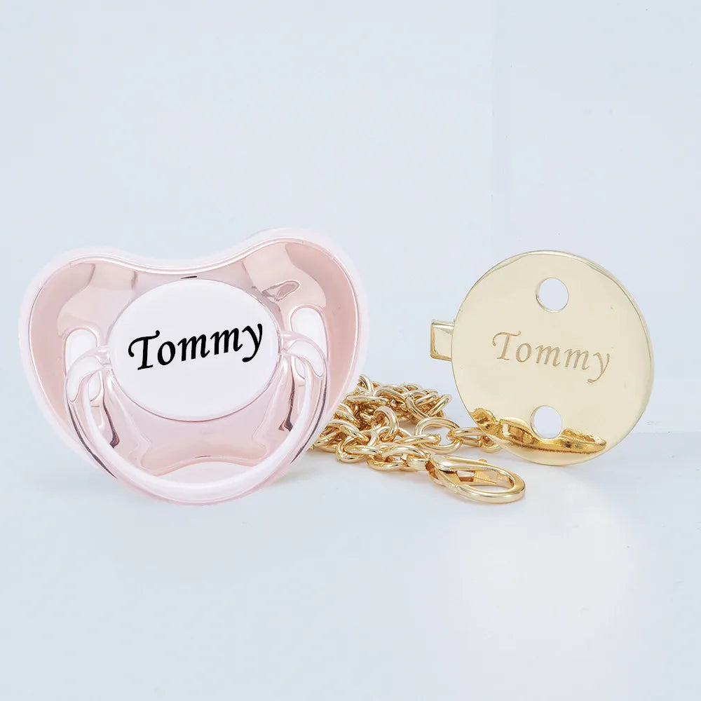 A pink silicone pacifier and a gold-tone clip with the name Tommy engraved on it.