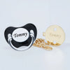 A set of personalized pacifier and clip featuring a black silicone body with a white disc and a gold-colored clip.