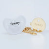 A clear silicone pacifier and a gold-colored clip with the name Tommy engraved on it.