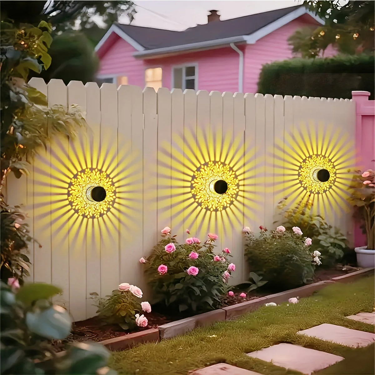 A pink house is illuminated by a white picket fence featuring solar peony wall lights shining bright yellow.