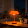 An orange mushroom-shaped LED table lamp with a slender stem sits on a wooden table in a dimly lit room.