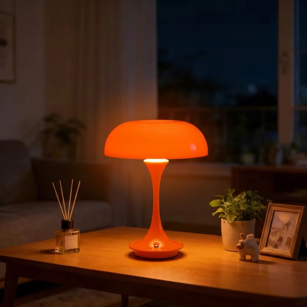 An orange mushroom-shaped LED table lamp with a slender stem sits on a wooden table in a dimly lit room.
