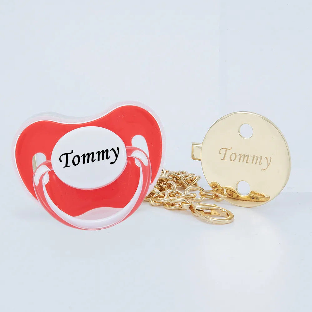 A red heart-shaped pacifier charm with the name Tommy and a gold-colored clip set.