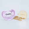 A pink silicone pacifier with the name Tommy and a gold-colored clip set.