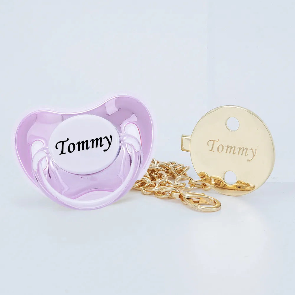 A pink silicone pacifier with the name Tommy and a gold-colored clip set.