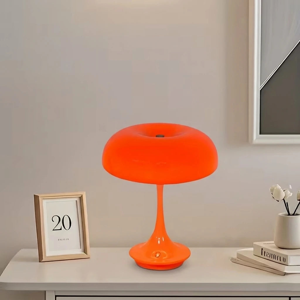 An orange mushroom-shaped LED table lamp with a slender stem sits on a white surface.