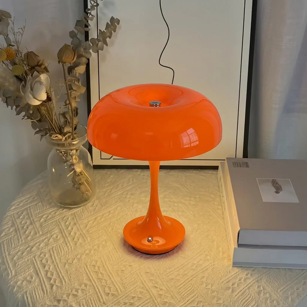 An orange mushroom-shaped LED table lamp with a slender stem sits on a white textured surface.