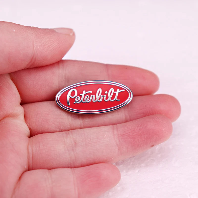 A hand holds a red and silver Peterbilt oval emblem enamel pin against a white background.