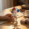 Eine Hand hält eine Kristall-Rosenfigur mit Glasvase, mit farbenfrohen Blütenblättern auf einem Holztable.