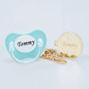 A light blue silicone pacifier and a gold-colored clip with the name Tommy engraved on it.