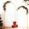 A gold metal wedding arch backdrop stand decorated with evergreen branches and red ornaments on a white wall.