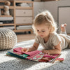 A toddler is interacting with a pink felt activity book on a white blanket.