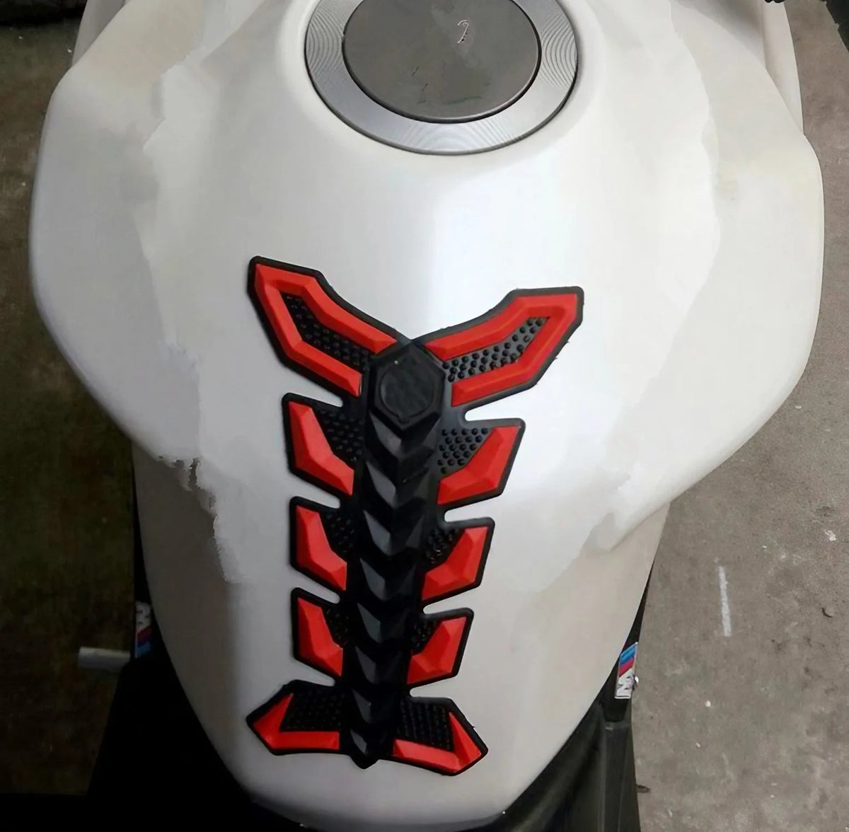 A close-up view of a white motorcycle tank pad protector with a black and red fish bone design.