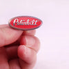 A hand holds a red Peterbilt oval emblem enamel pin against a white background.