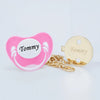 A pink silicone pacifier with the name Tommy and a gold-colored clip set.