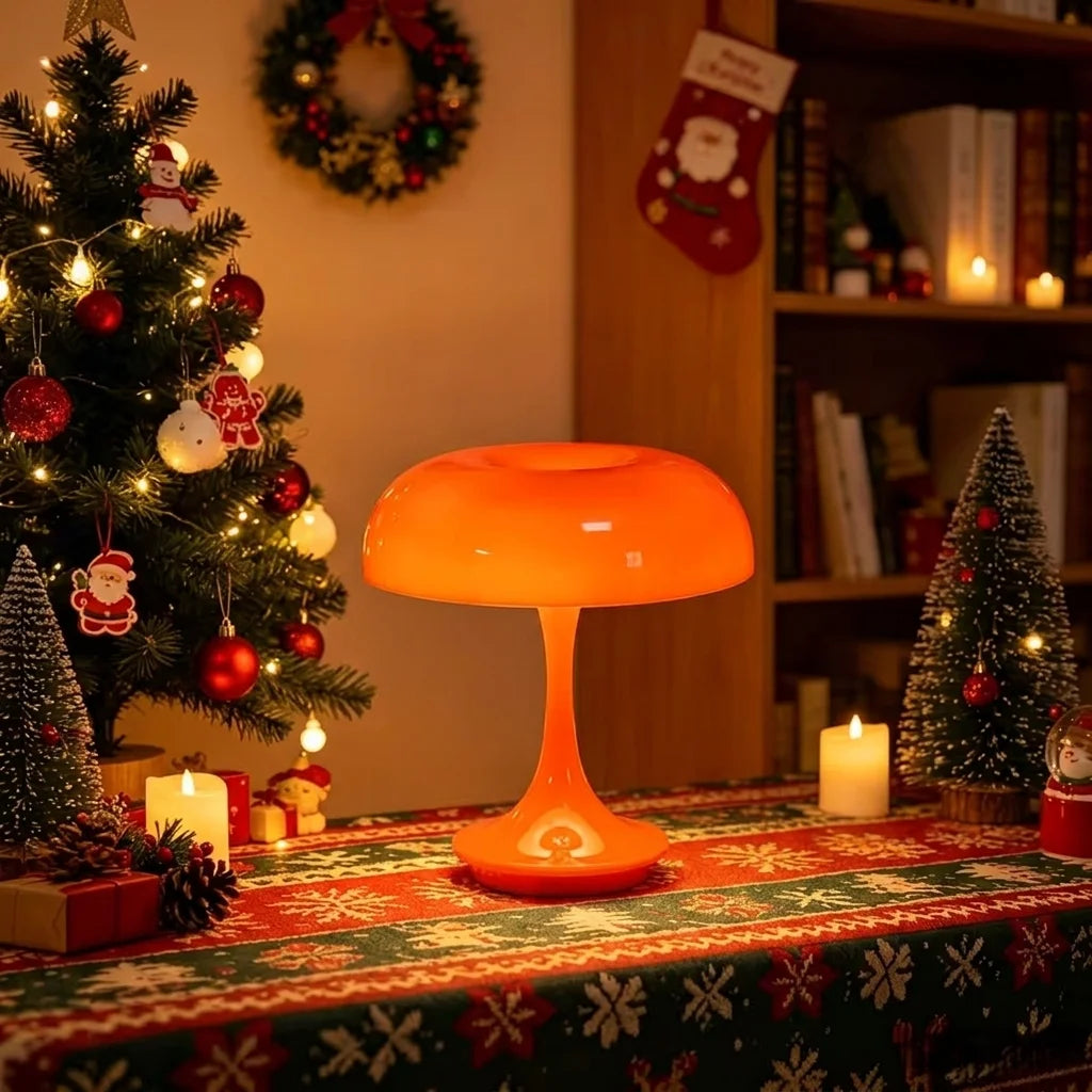 An orange mushroom-shaped LED table lamp sits on a festive red and green tablecloth near a decorated Christmas tree.