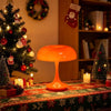 An orange mushroom-shaped LED table lamp sits on a festive red and green tablecloth near a decorated Christmas tree.
