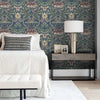 A bedroom wall featuring blue vintage damask floral wallpaper, a bedside table with a lamp, and white bedding.