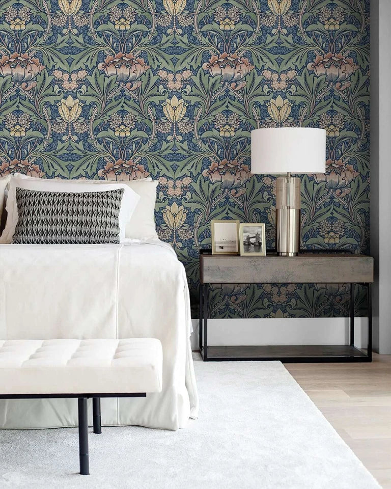 A bedroom wall featuring blue vintage damask floral wallpaper, a bedside table with a lamp, and white bedding.