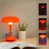 An orange mushroom-shaped LED table lamp with a white base sits on a wooden surface next to an open book.