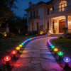 A stone pathway illuminated by a line of colorful solar LED garden lights leading toward a large house at dusk.