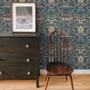 A dark wooden dresser with a framed photo stands next to a wooden chair against blue vintage damask floral wallpaper.