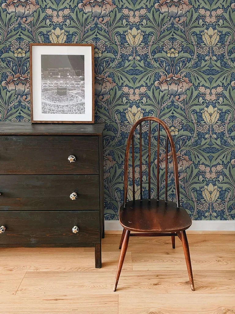 A dark wooden dresser with a framed photo stands next to a wooden chair against blue vintage damask floral wallpaper.