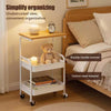 A white 3-tier rolling utility cart with a wooden top, holding a lamp and a teddy bear, next to a wooden bed frame.