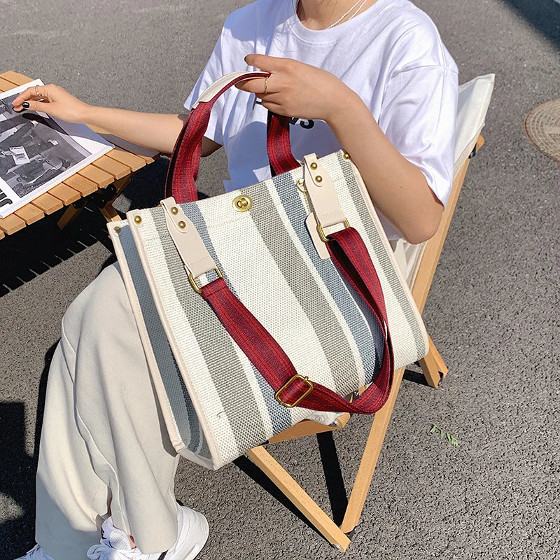 A person holds a beige and striped canvas crossbody tote bag with red straps while sitting outdoors.