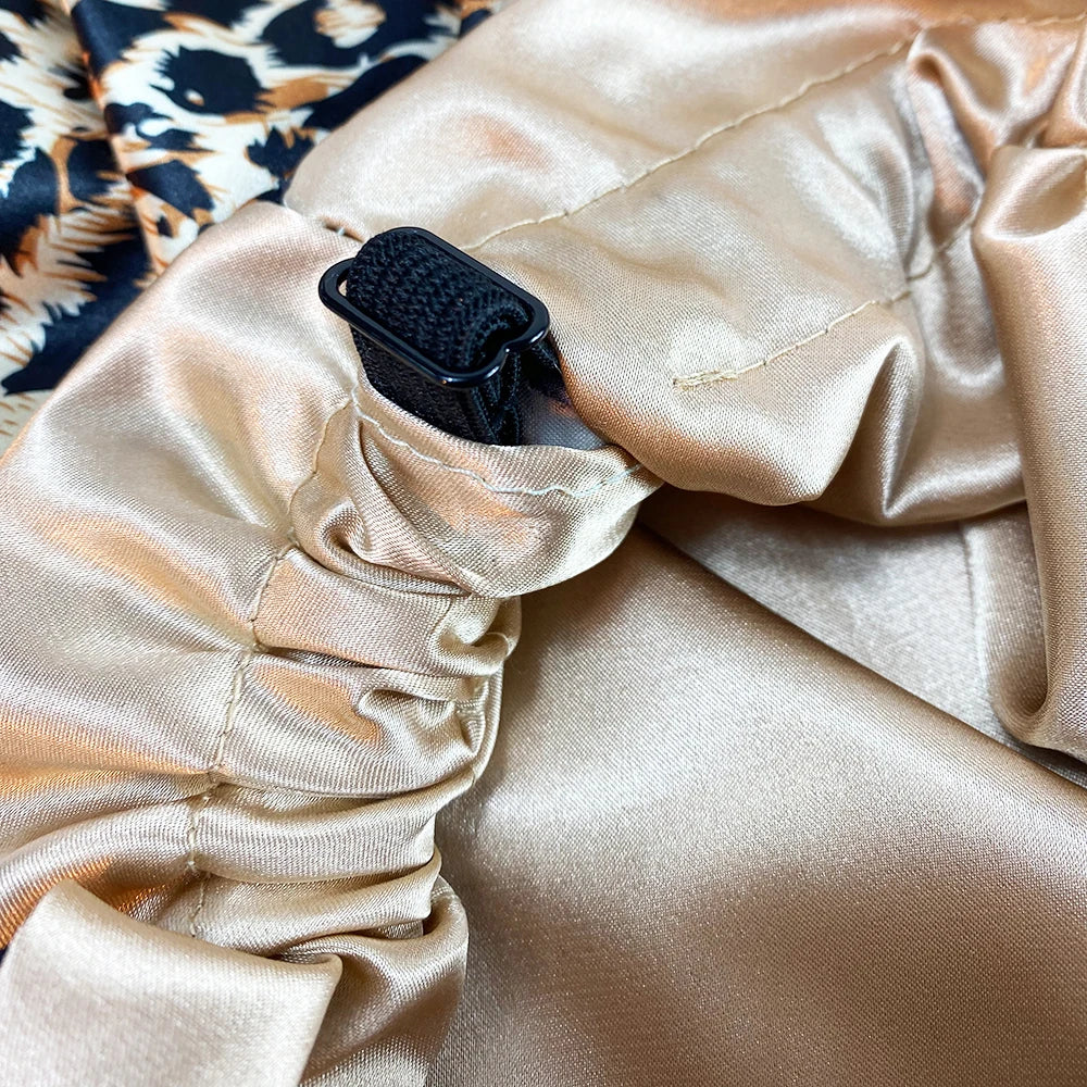A close-up of a double-layer satin hair bonnet with an adjustable black band, shown against a smooth beige satin fabric.
